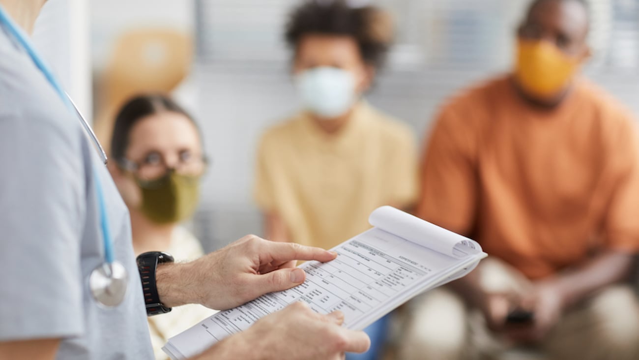 A male physician holds a clipboard in a waiting room with patients wearing masks.