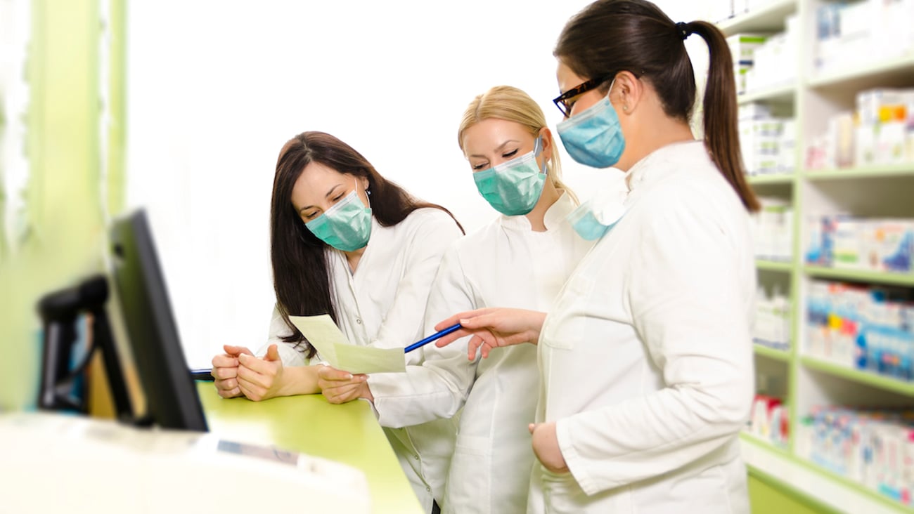 Three female pharmacists wearing lab coats and masks stand behind a pharmacy counter looking at documents together.