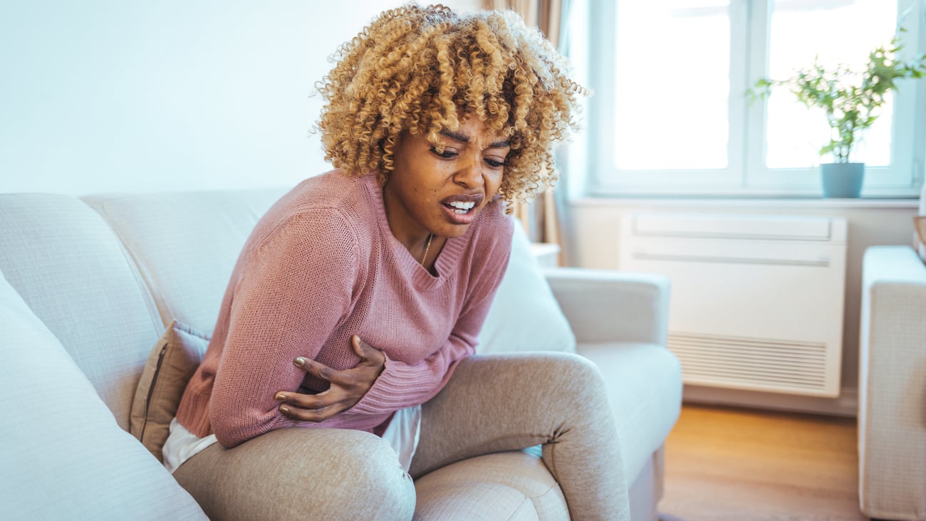 Black woman with light hair wearing pink sweater holds stomach in pain and grimaces