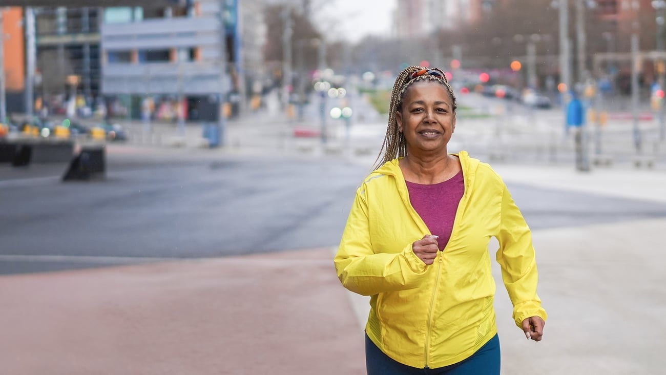 Black woman running toward the camera, smiling in bright yellow jacket; urban street in background