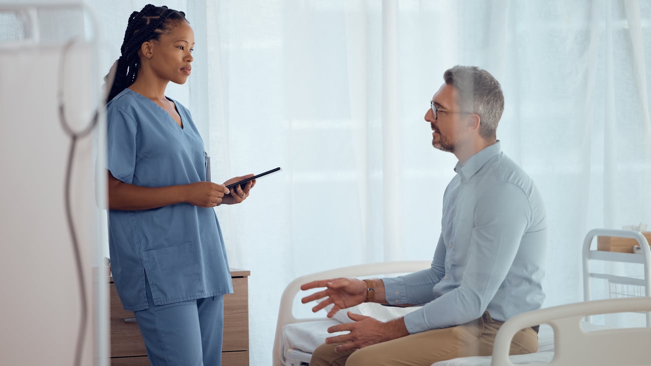 Young Black woman doctor in blue scrubs speaking with older white male patient sitting on the exam table