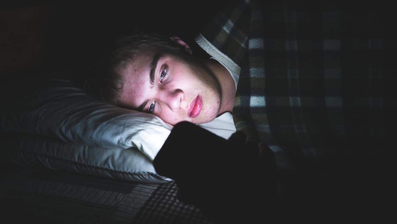 serious face of teen boy in dark in bed looking at his phone, with phone's light shining on his face