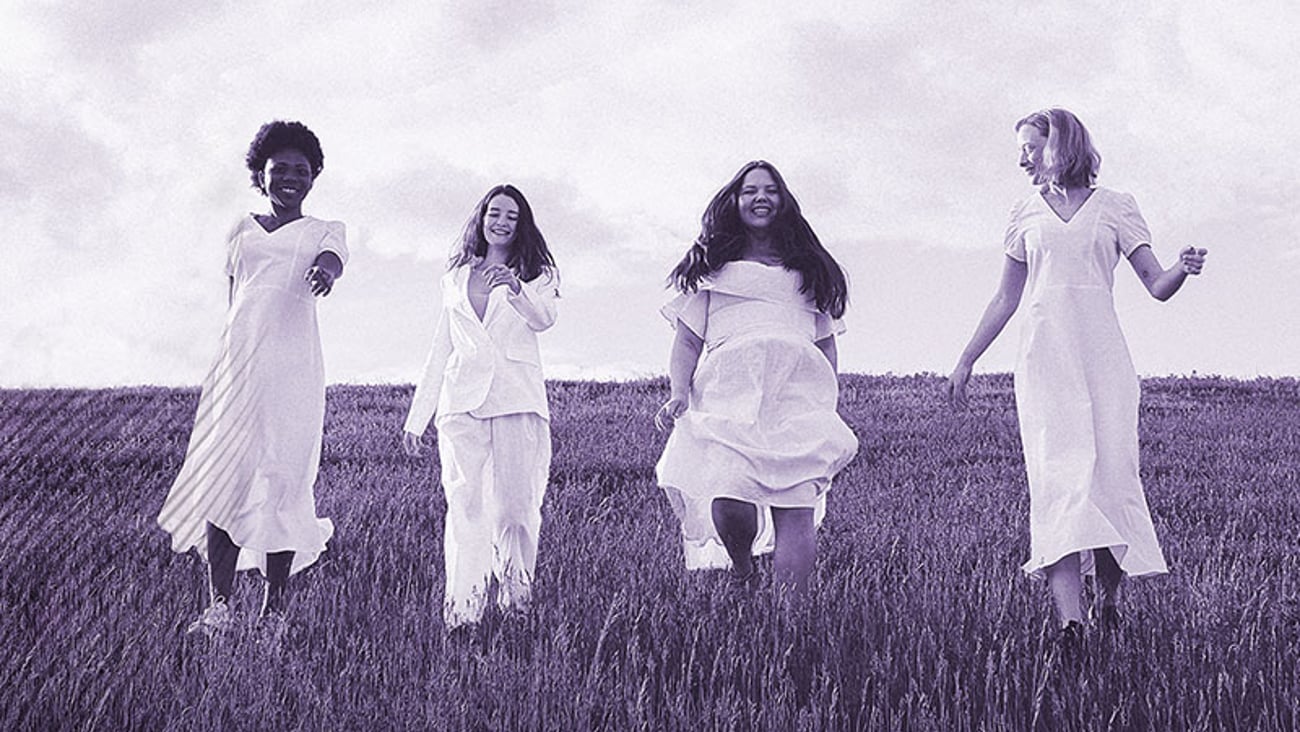 four women of various ages wearing white dresses walk across an open field