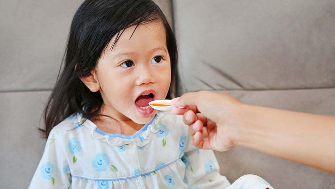 young female child sits on a sofa with her mouth open, about to receive a dose of medicine on a spoon