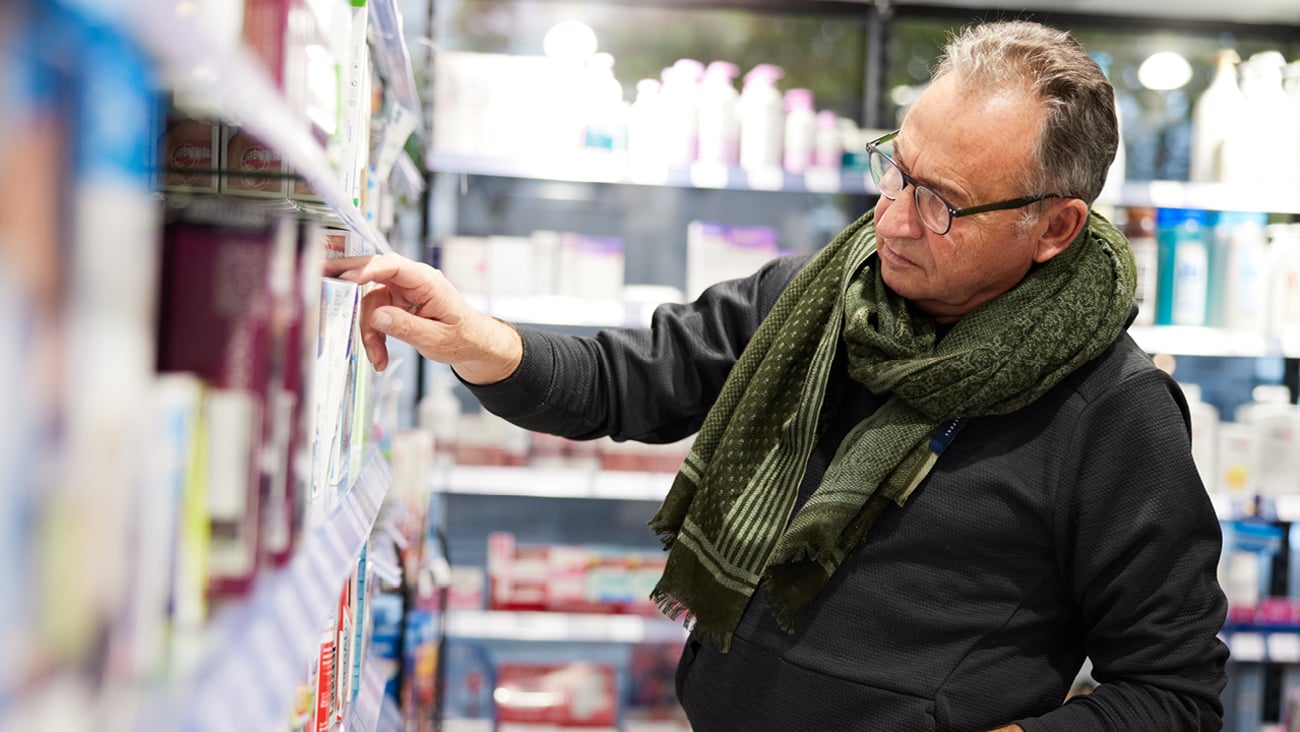 Older male customer in stylish green scarf looking quizzically at natural health products on a pharmacy shelf