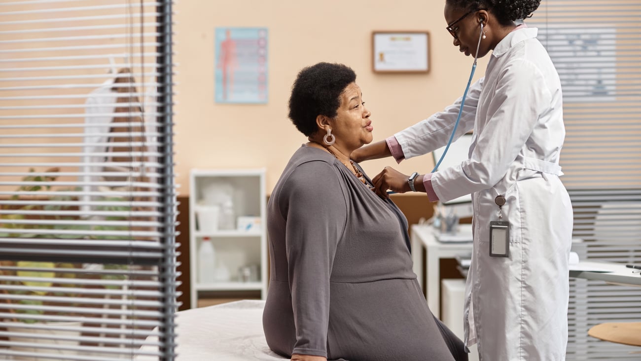 Side view of senior Black woman sitting on therapy bed in medical office breathing out while female health practitioner listening to lung sounds