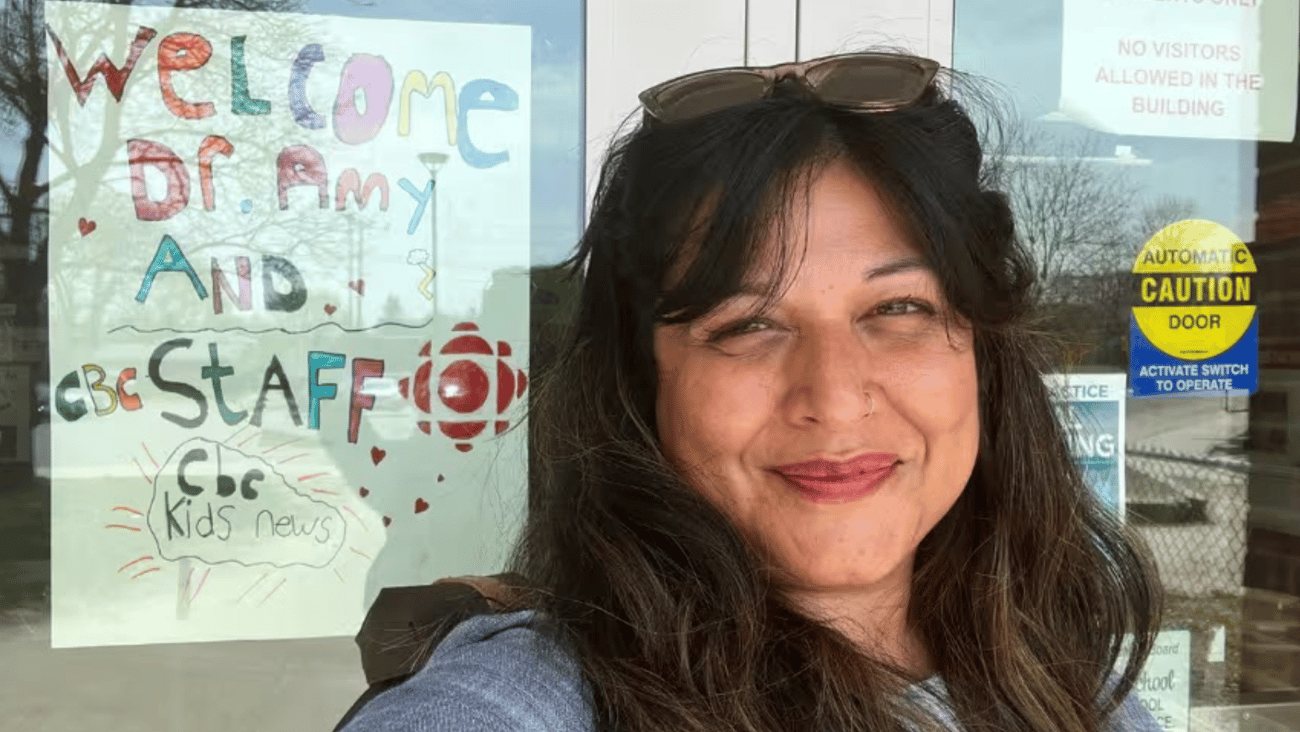Dr. Amy Gajaria, a brown-skinned woman with long brown hair, smiling in front of a set of glass doors
