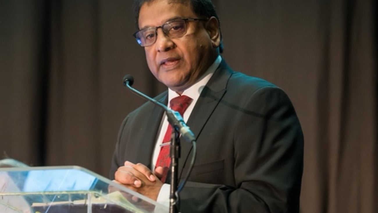 Sham Moodley, a brown-skinned man in a black suit and glasses giving a speech at a podium