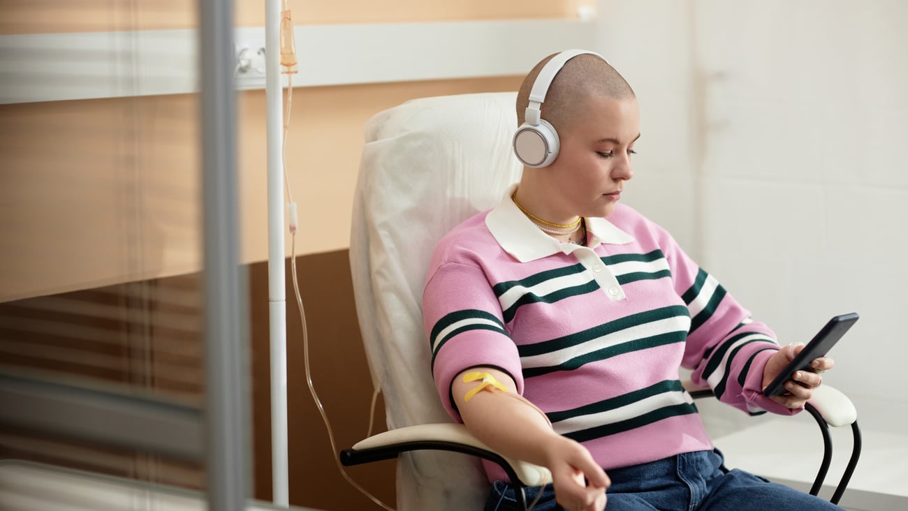 Young woman, bald from chemotherapy treatment, calmly looking at her phone while receiving chemo treatment