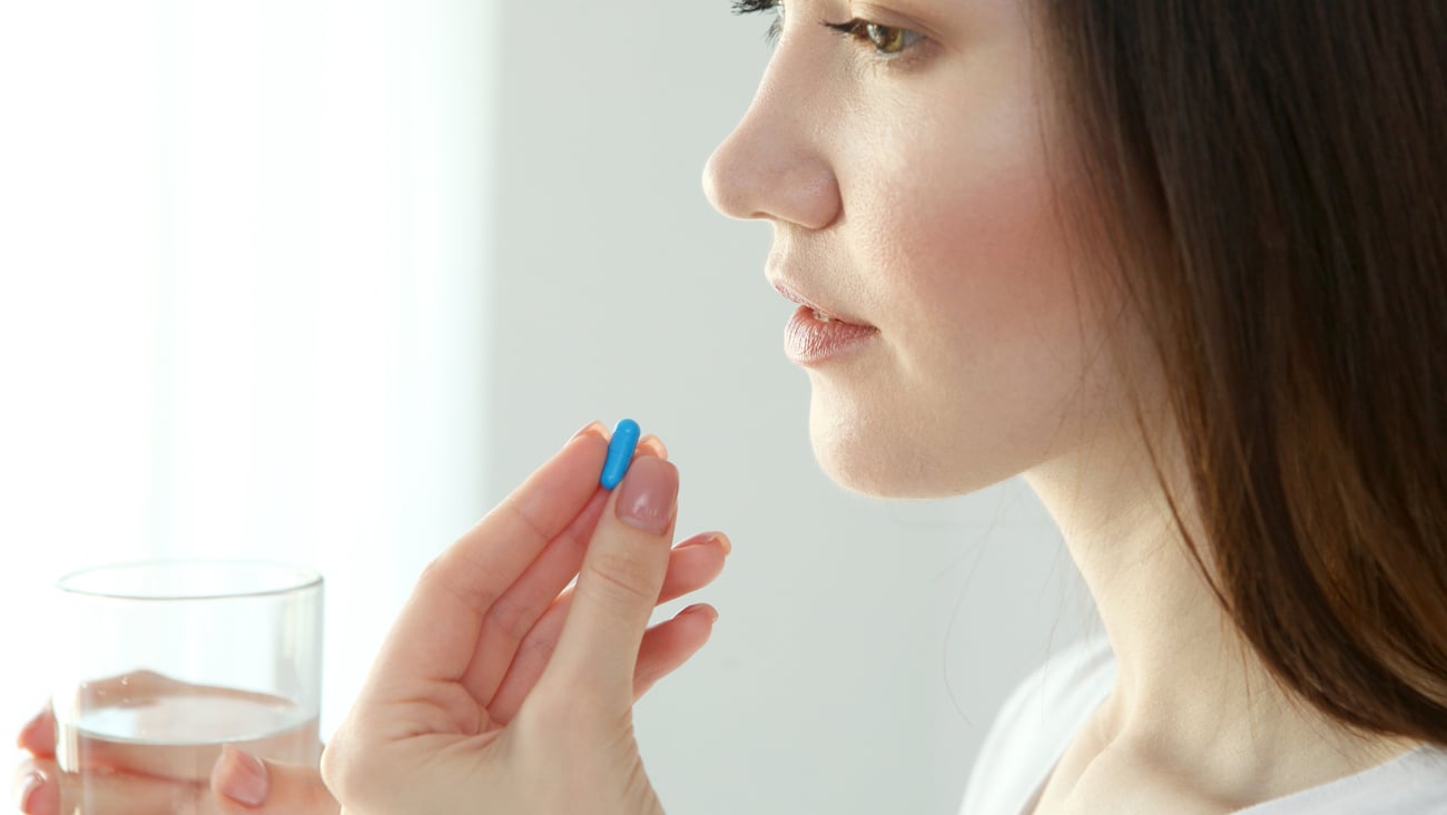 Young white woman taking blue pill with glass of water, looking pensive