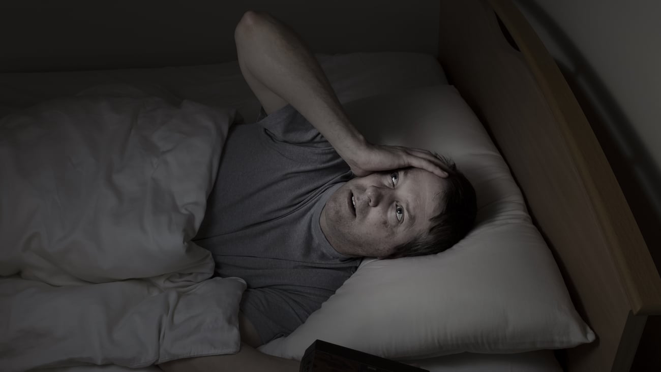 man in darkness staring at ceiling looking troubled, hand on side of face