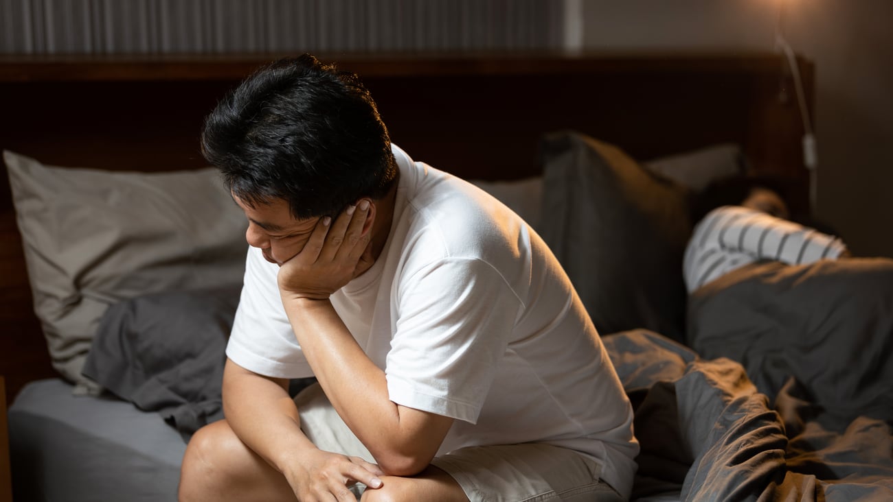Young man sitting dejected on the side of his bed while his partner sleeps