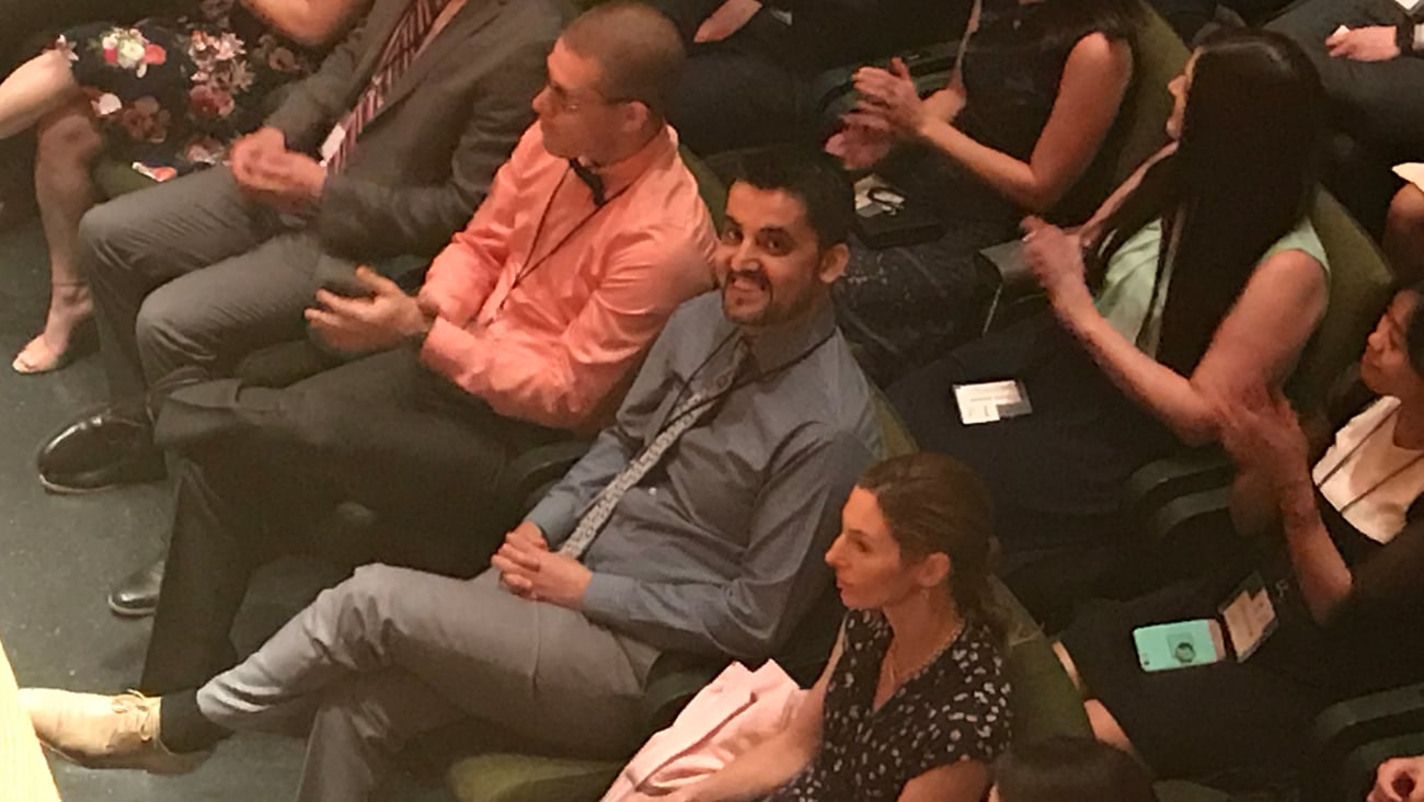 A man sitting in a large audience, smiling up at someone in the balcony