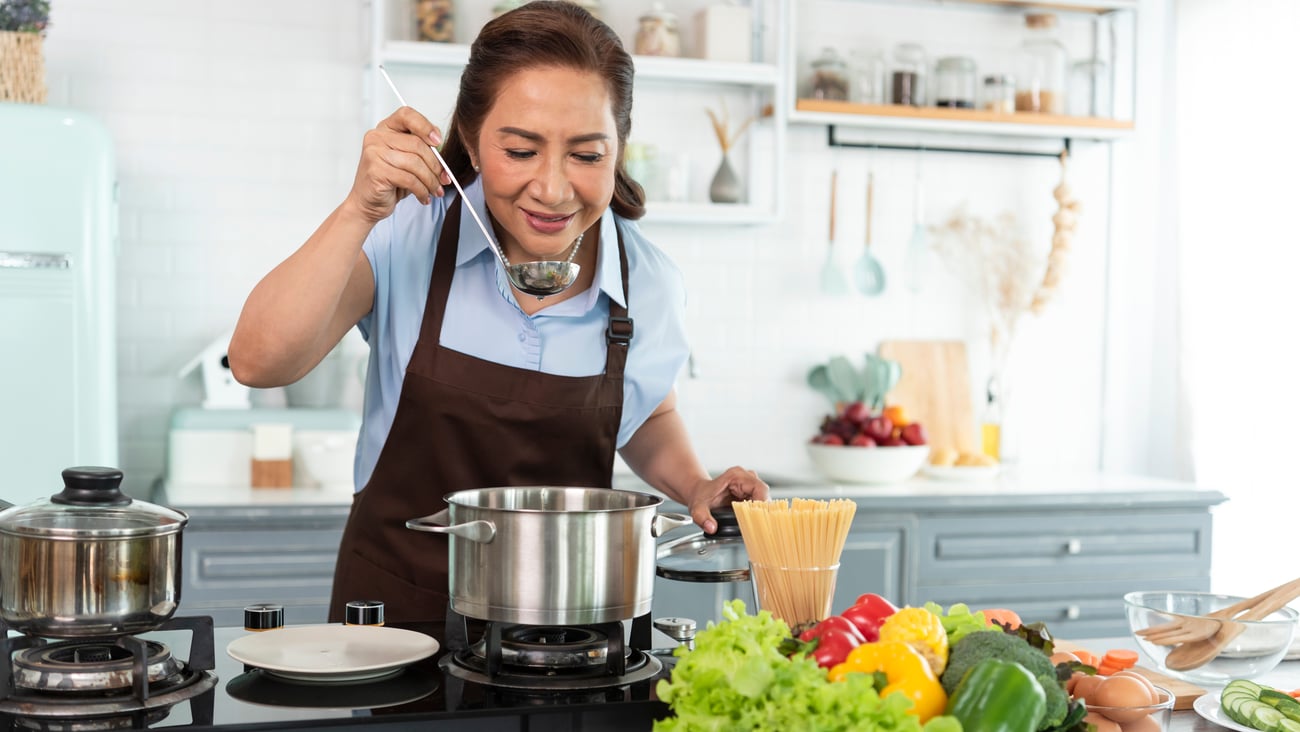 Happy smiling asian older woman in apron is tasting and smell food while cooking in kitchen.