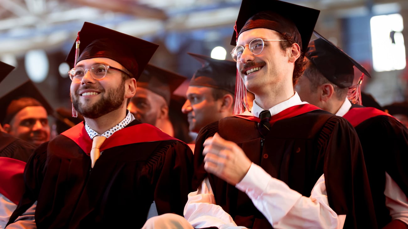 A man with a beard and a man with a mustache sit smiling beside each other wearing academic robes at a convocation ceremony.