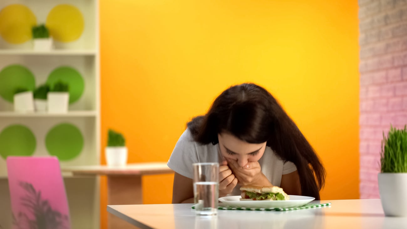 Picture of girl/young woman at table in front of plate, folded over with hand on mouth as if gagging or nauseated.