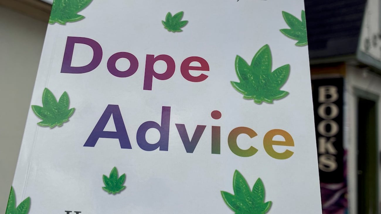 a copy of the paperback book Dope Advice is held up against a cloudy sky with a cute book store in the background