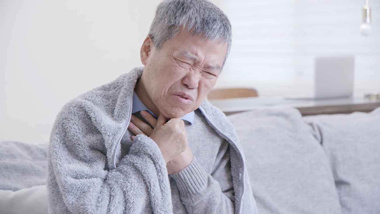 An elderly asian man in grey sweater clutches his sore throat