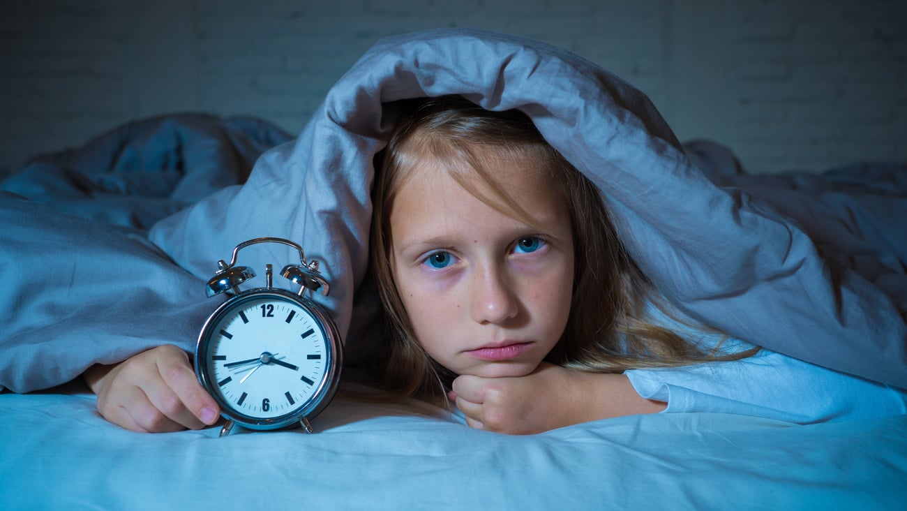 Young girl unable to sleep, under her covers peeping out with an alarm clock