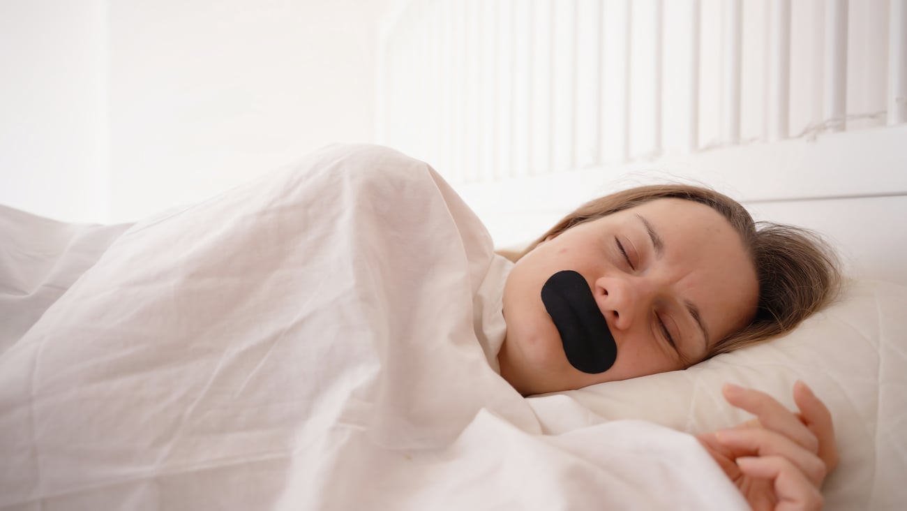Woman sleeping on side under white blanket with black mouth tape in bright bedroom