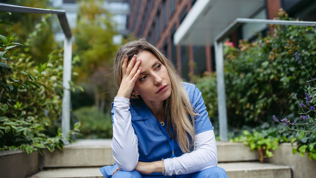 Young woman doctor sitting on the steps outside a hospital with her head in her hand in frustration
