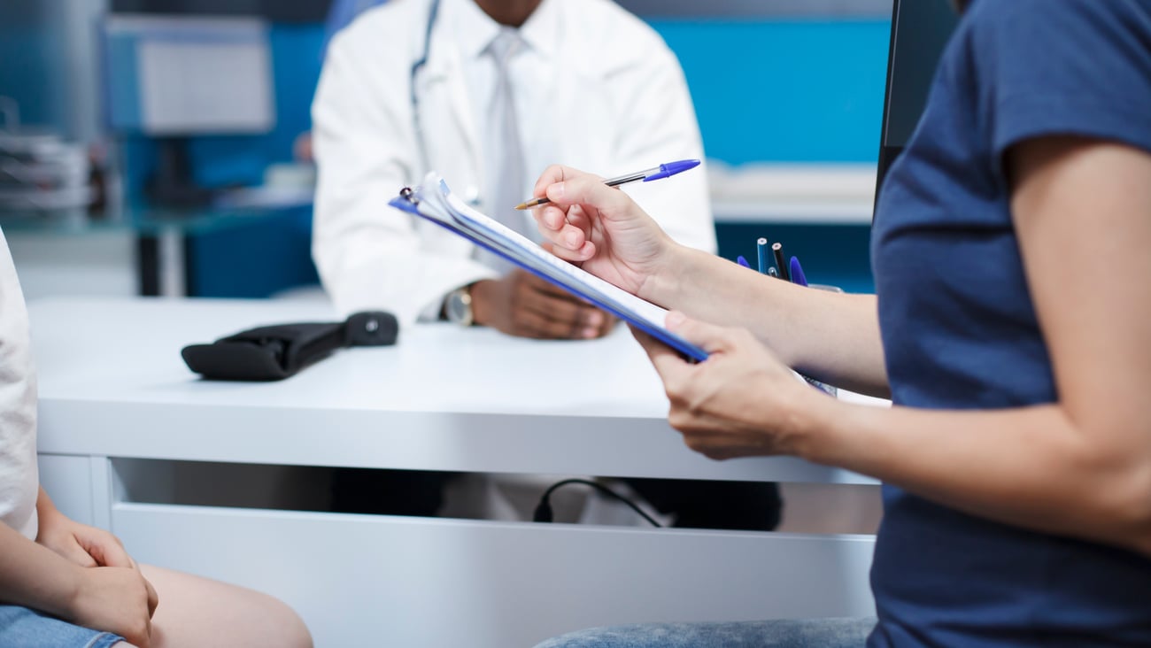 A young woman healthcare worker asking a young patient questions with another doctor in the background