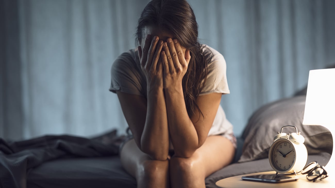 Young woman sitting on the side of her bed with her head in her hands
