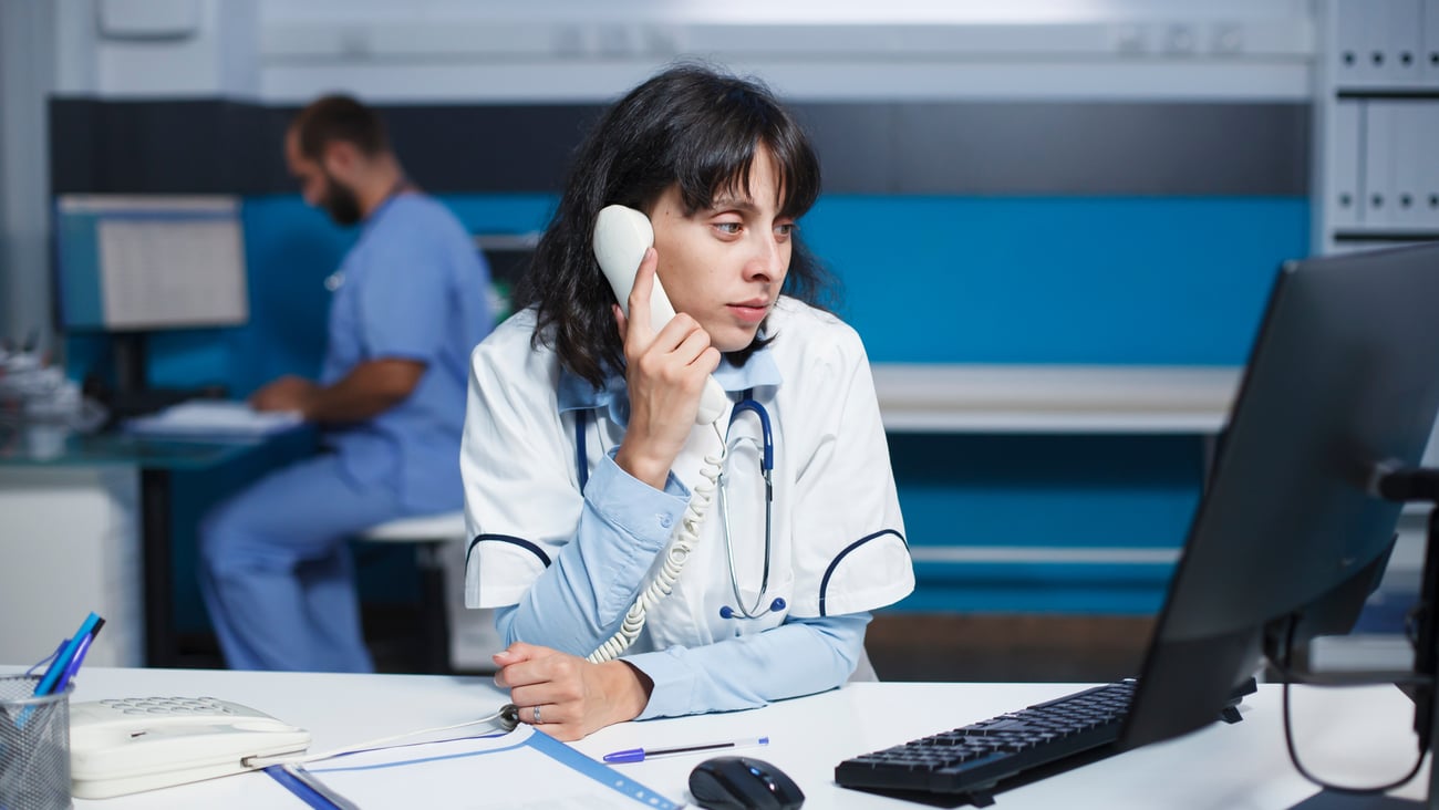 A doctor making a phone call in front of a desk at an office.