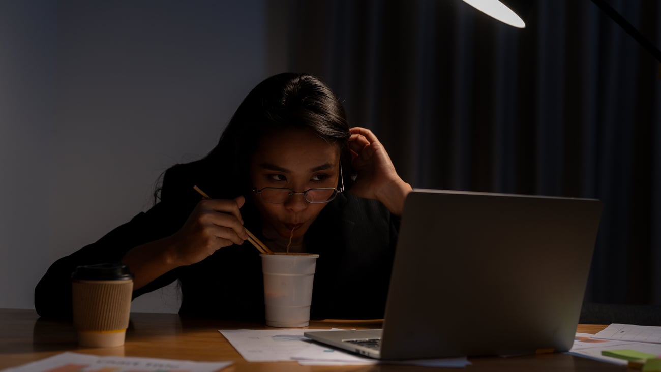 Young woman working late eating noodles while looking at her laptop
