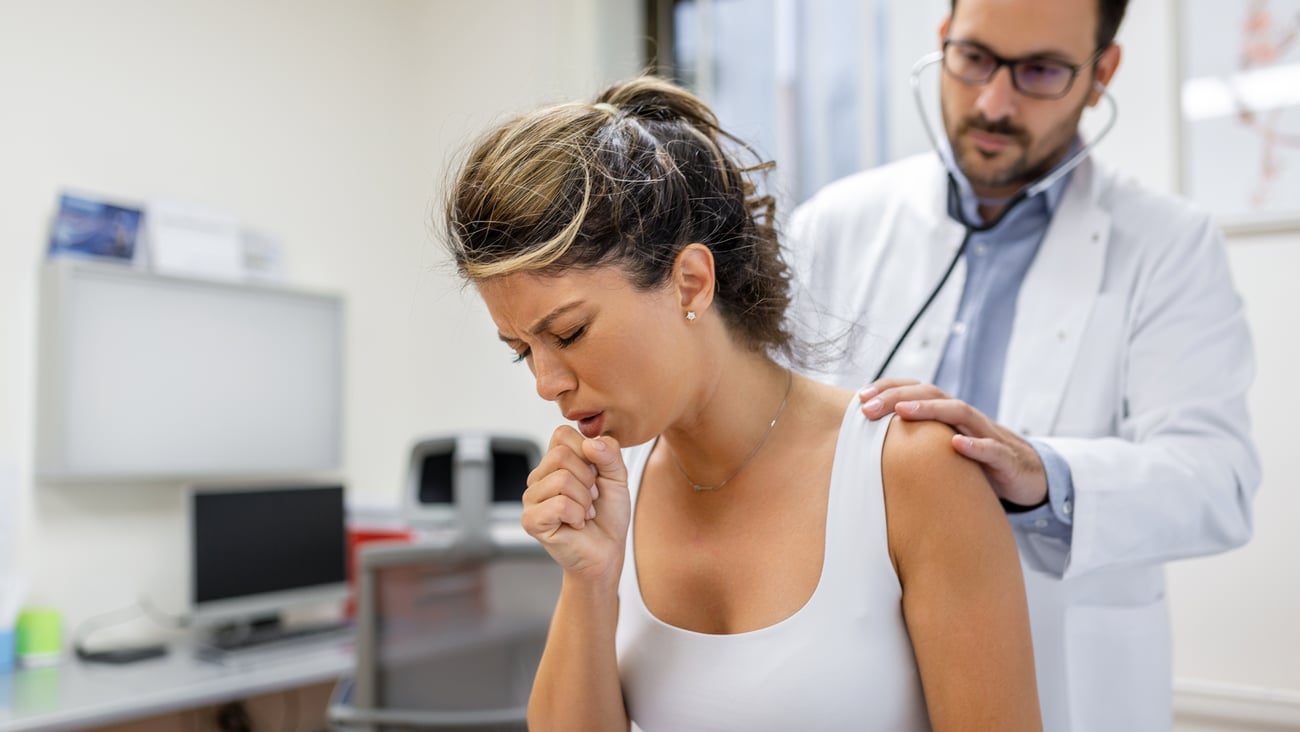 Young woman coughing with a doctor listening to her lungs behind her