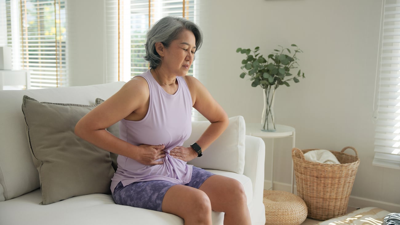 Older Asian woman in a purple shirt holding her stomach in discomfort