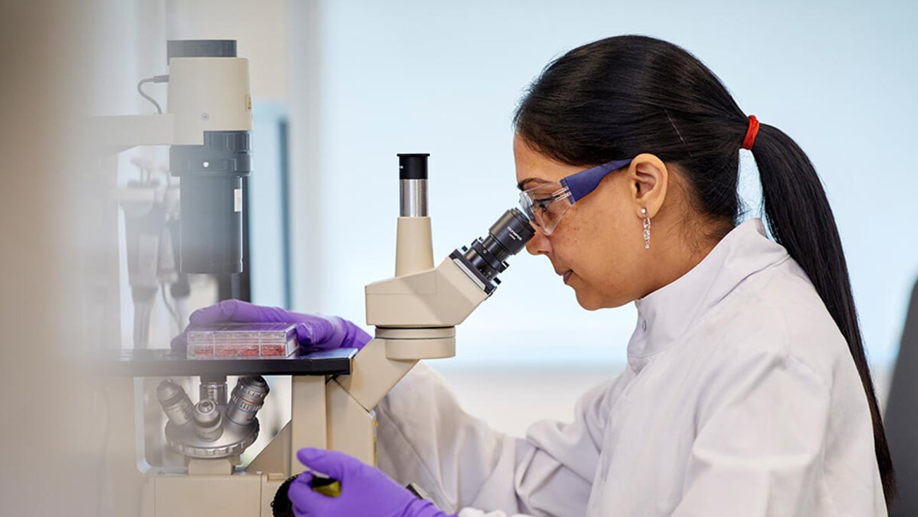 A female scientist in a white lab coat and safety glasses intently examining a sample through a professional laboratory microscope.