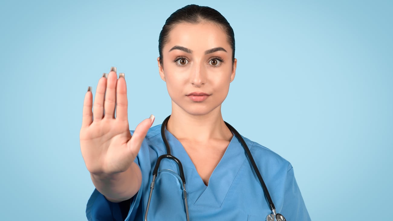 Composed female medical professional in blue scrubs makes firm stop gesture, conveying authority and importance of health protocols against serene blue backdrop