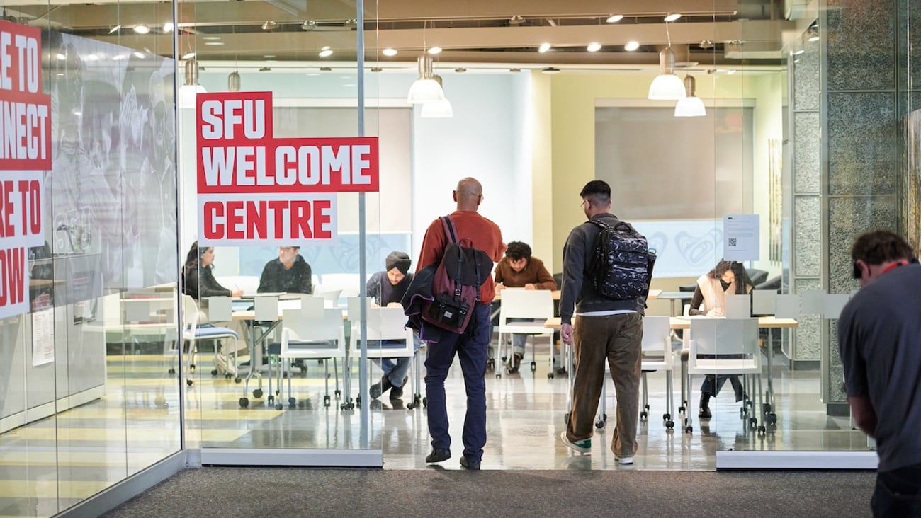 Two young people walk through a glass door beside a glass wall with the words SFU welcome centre