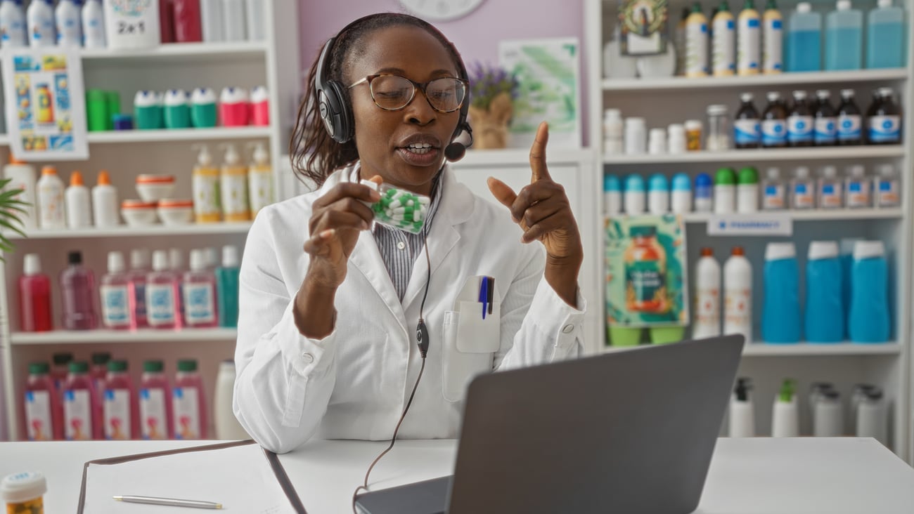 Female pharmacist in white coat with headset advising medication to customer via laptop in drugstore