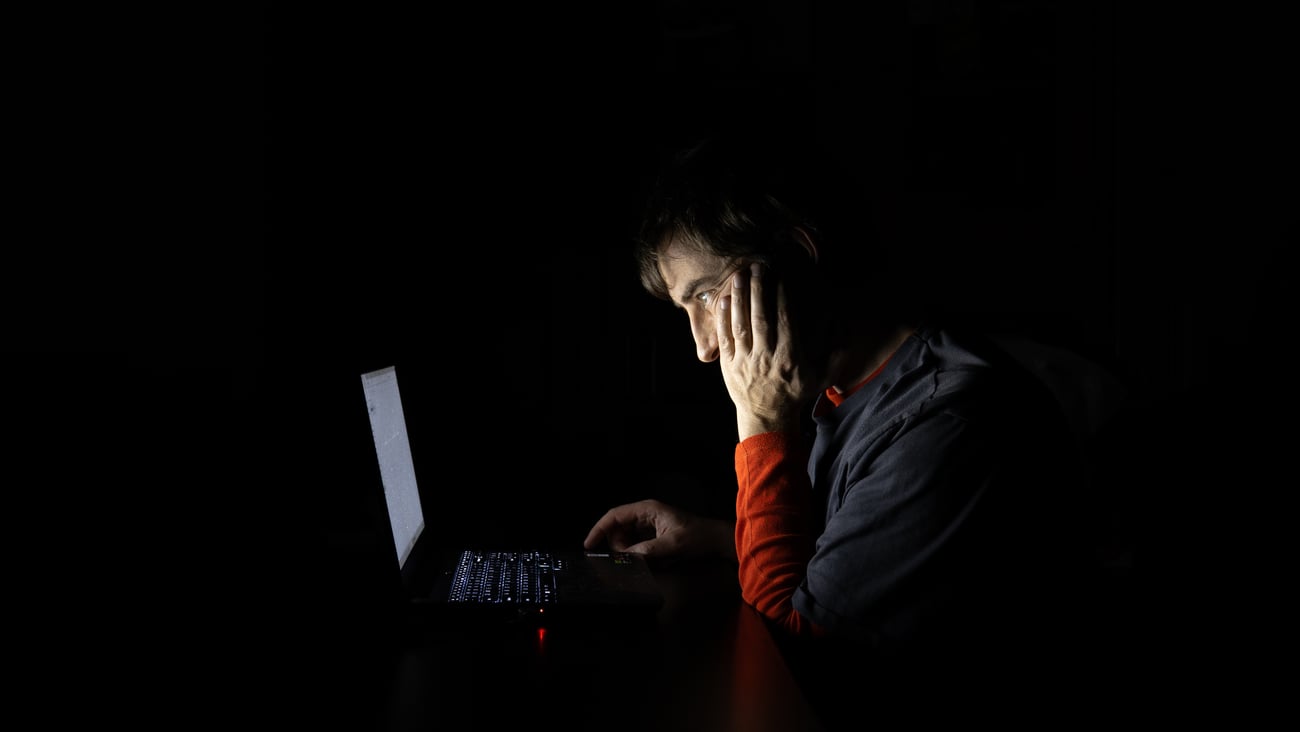 Side profile of an exhausted man resting his head on his hand while looking at a glowing laptop screen in a completely dark room