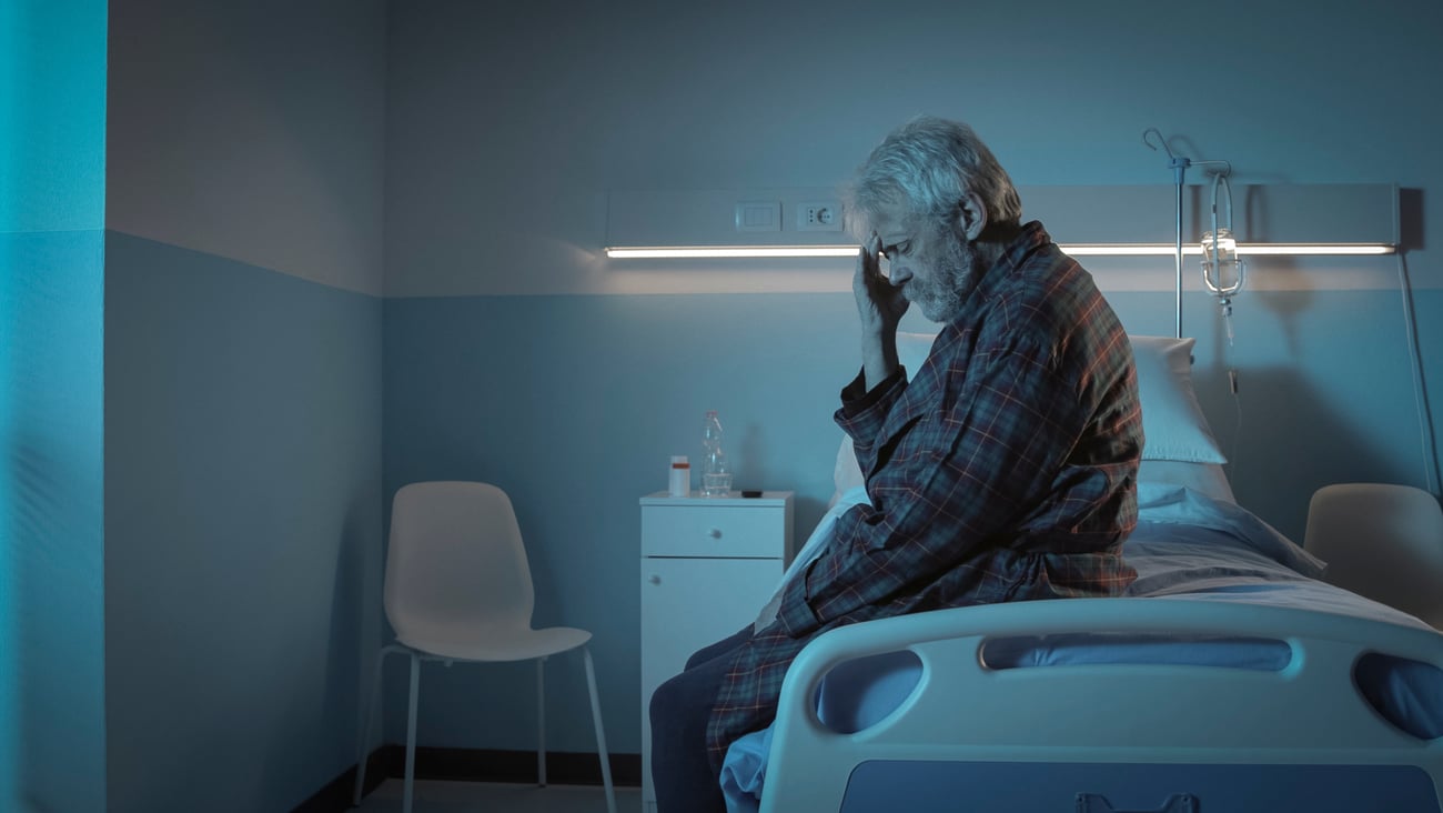 A senior with his hand on his head while sitting on a hospital bed.
