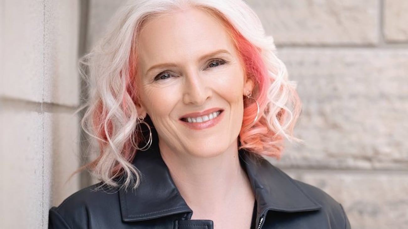 A white physician woman with blond and pink hair smiles at the camera in a leather jacket.