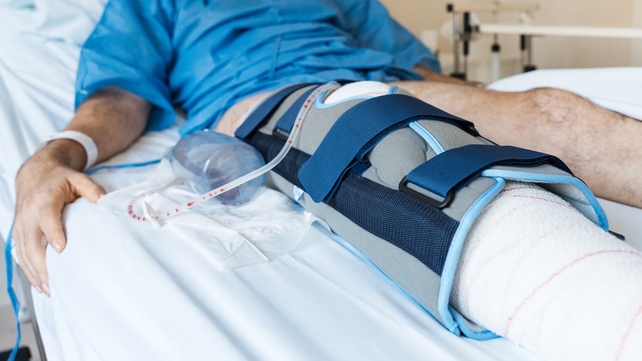 Male patient lying in hospital bed after leg surgery, wearing knee brace and cast with medical drain attached during recovery