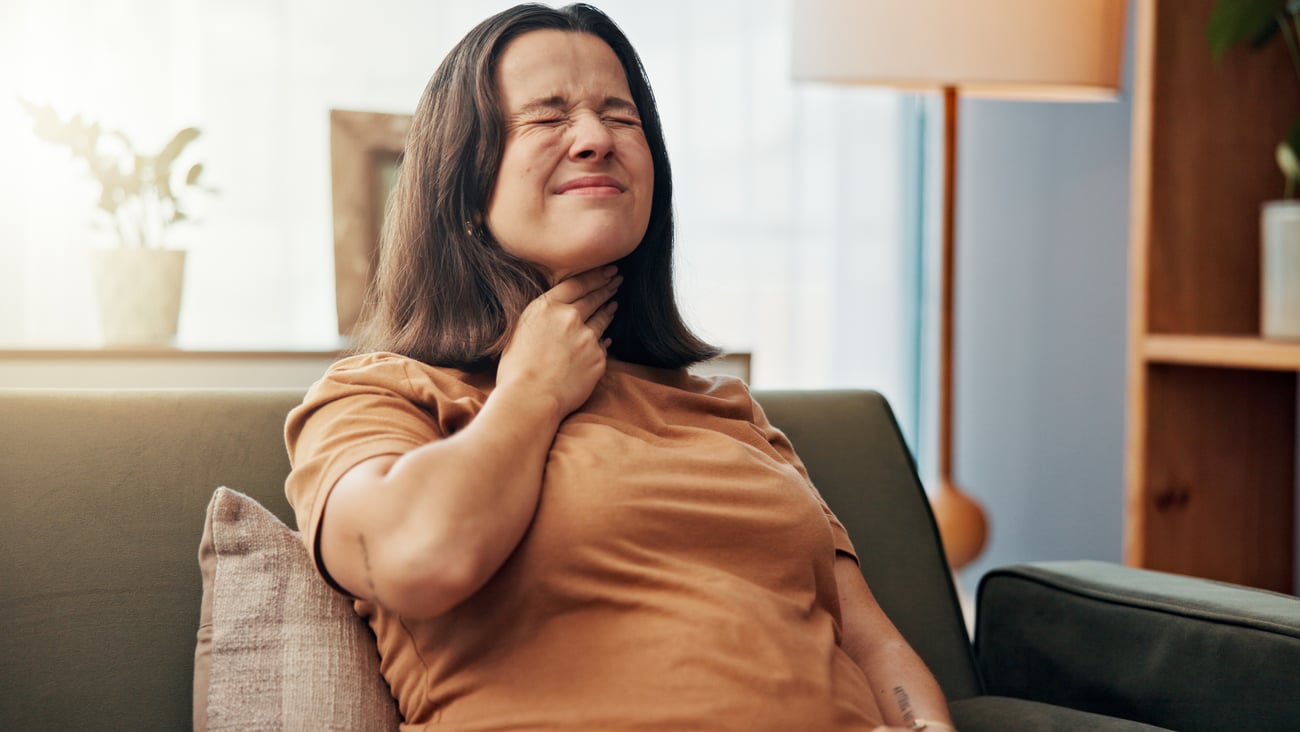 Young white woman holding her throat in discomfort at home on the couch