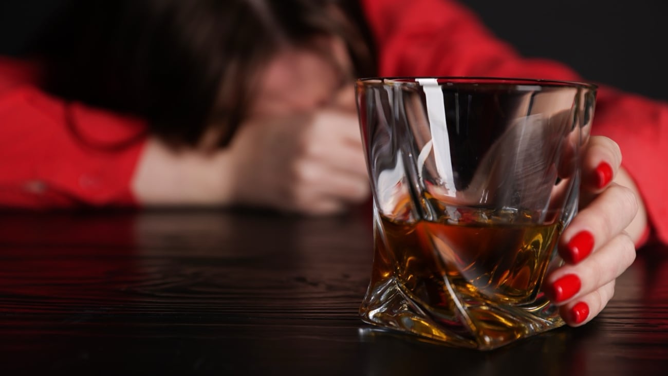 A woman hunched over with a glass of whisky in the foreground.