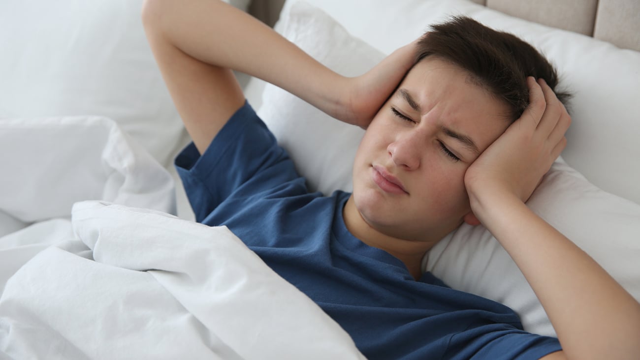 A young teenage boy holding his head in bed with a migraine