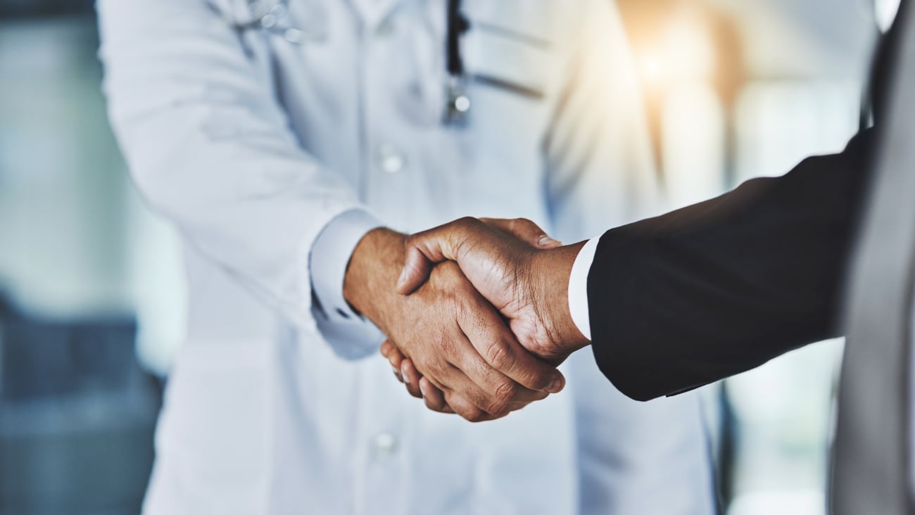 A doctor wearing a white coat shakes hands with a person in a suit.