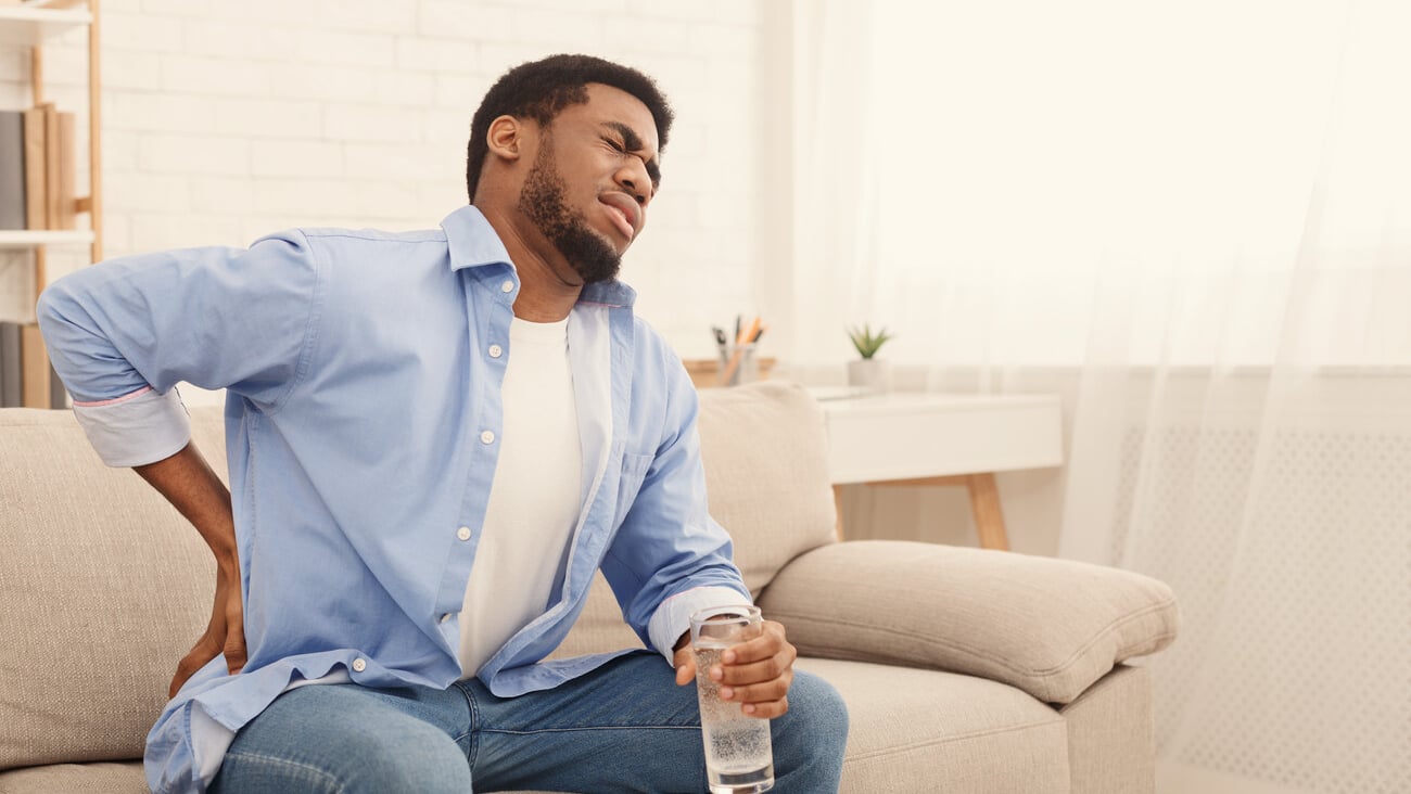 Young african-american man with back pain, pressing on hip with painful expression, sitting on sofa at home with glass of water, copy space; Shutterstock ID 1286261095