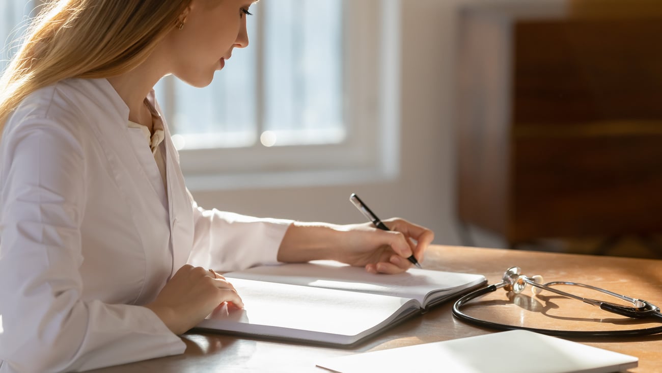 Focused millennial nurse or family doctor therapist general practitioner involved in paperwork, sitting at table in clinic office. Skilled professional physician recording notes in registry book.; Shutterstock ID 1917304301