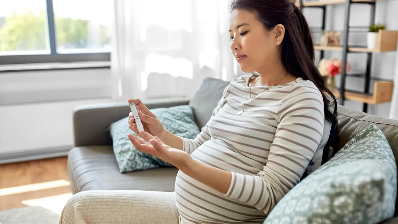 pregnancy, health and glycemia concept - pregnant asian woman checking blood sugar level with glucometer and lancing device at home; Shutterstock ID 2011679222