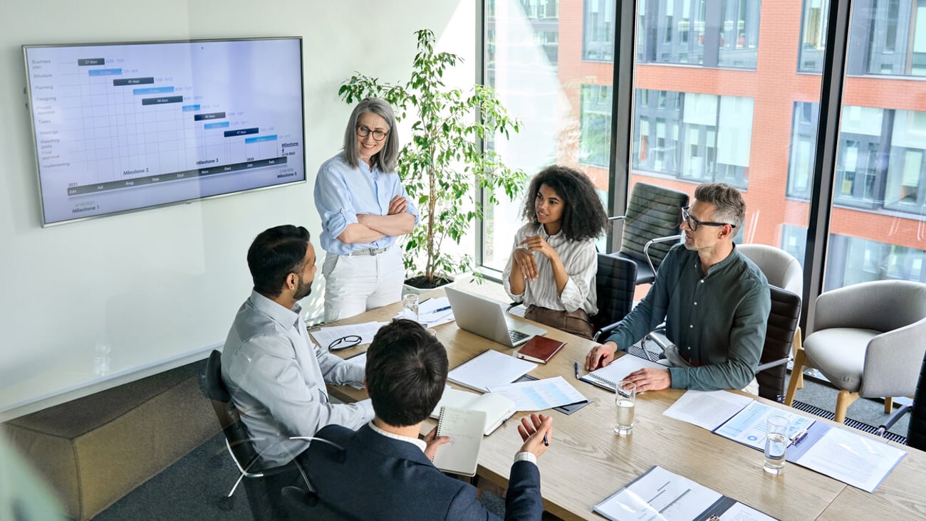 Senior female ceo and multicultural business people discussing company presentation at boardroom table. Diverse corporate team working together in modern meeting room office. Top view through glass; Shutterstock ID 2021639270