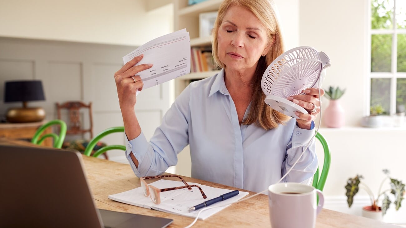 Menopausal Mature Woman Having Hot Flush At Home Cooling Herself With Fan Connected To Laptop; Shutterstock ID 2180443267