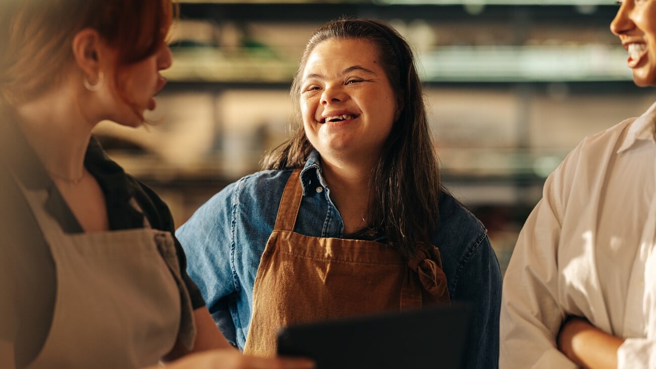 Retail worker with Down syndrome smiling happily while standing in a staff meeting in a grocery store. Group of diverse women working together in an all-female small business.; Shutterstock ID 2206851051