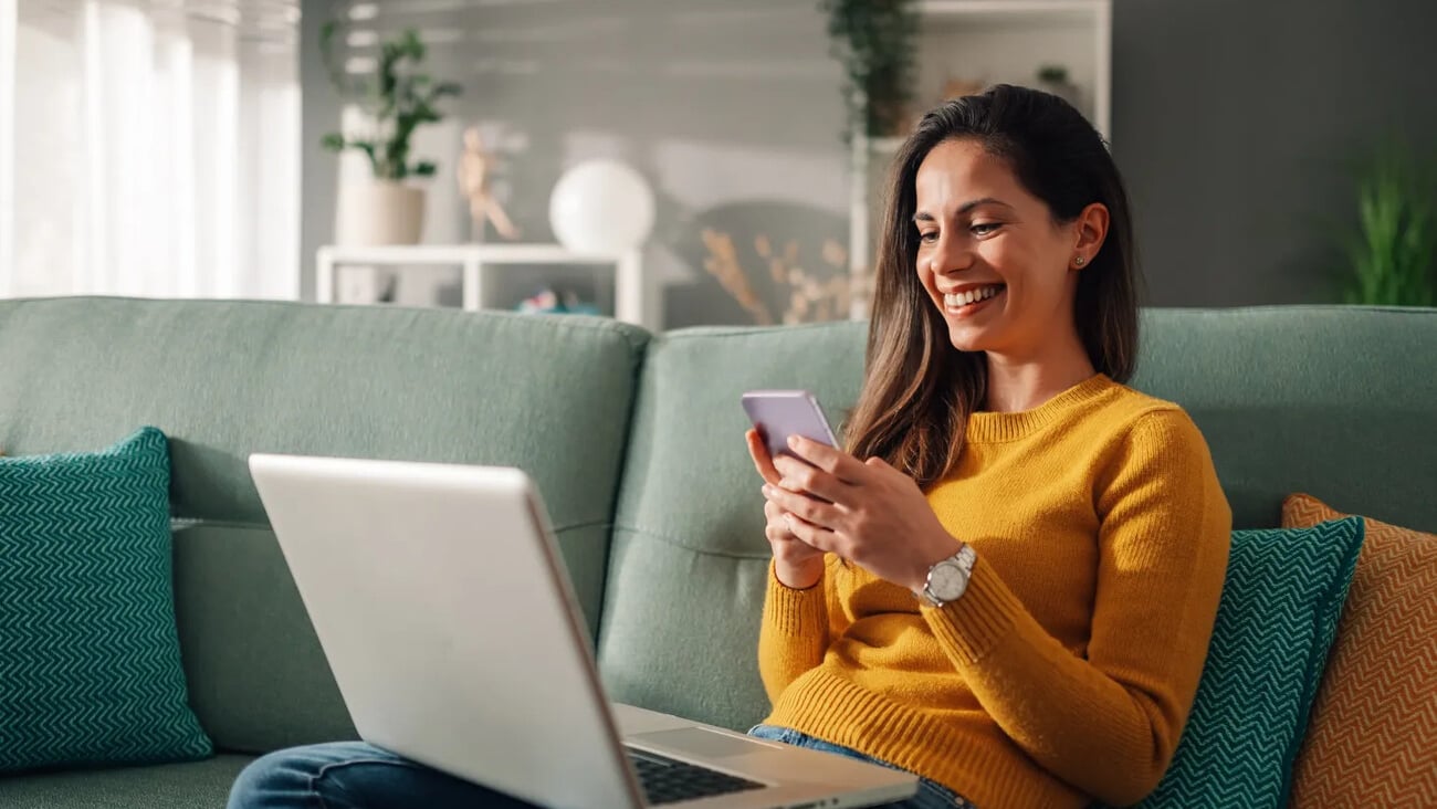 Portrait of a woman using laptop and a phone and checking email or news online while sitting on sofa at home. Searching for friends in internet social networks or working on computer. Copy space.; Shutterstock ID 2418478753